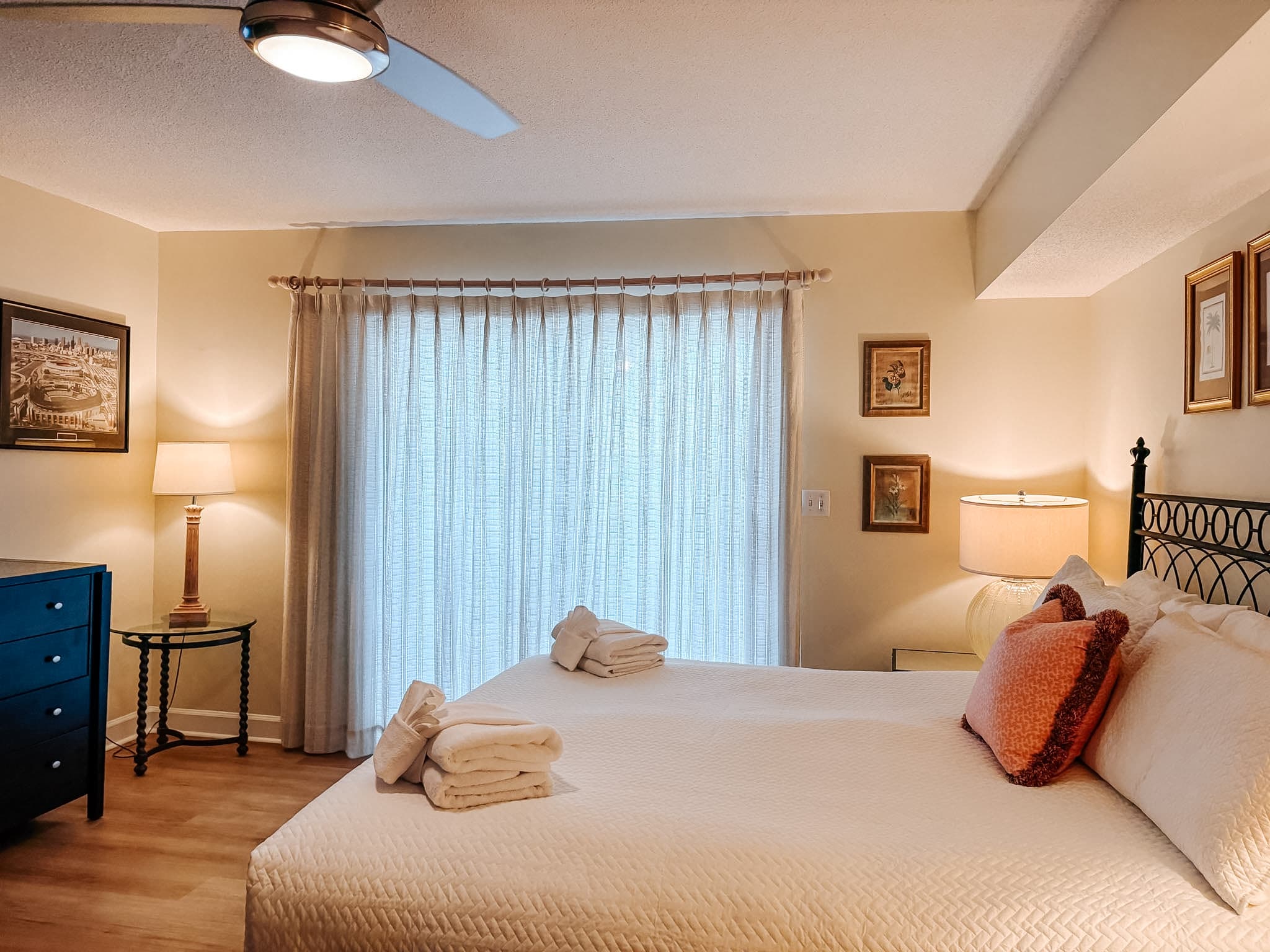 Cozy bedroom featuring white linens, folded towels, warm lamps, and large sheer curtains.