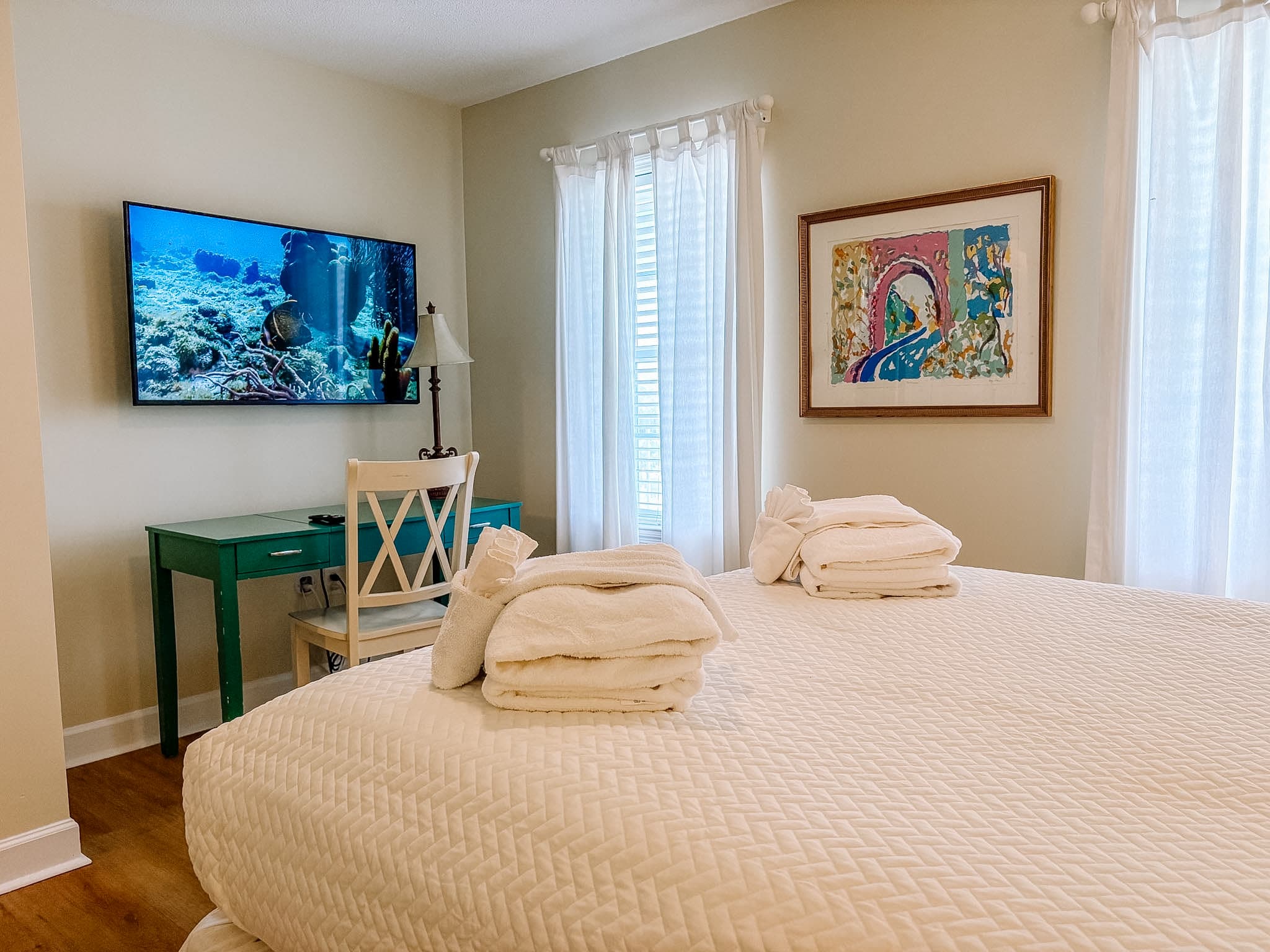 Bright bedroom with white quilted bed, green desk, wall-mounted TV, and framed abstract painting.