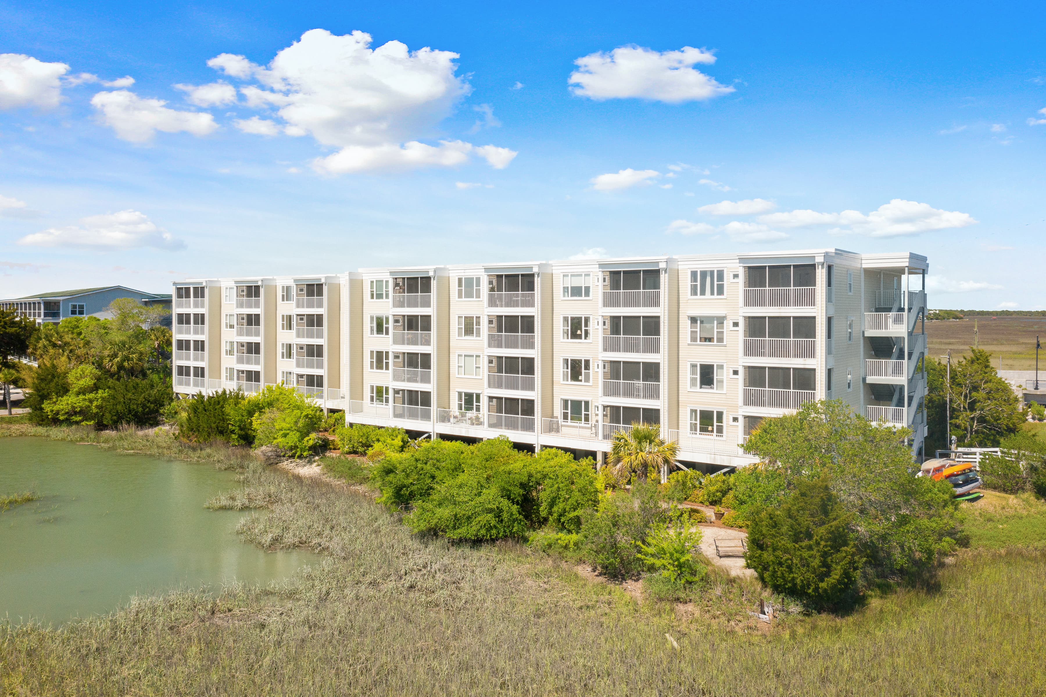 Multi-story white condo building with balconies overlooking a green coastal marsh under blue skies.